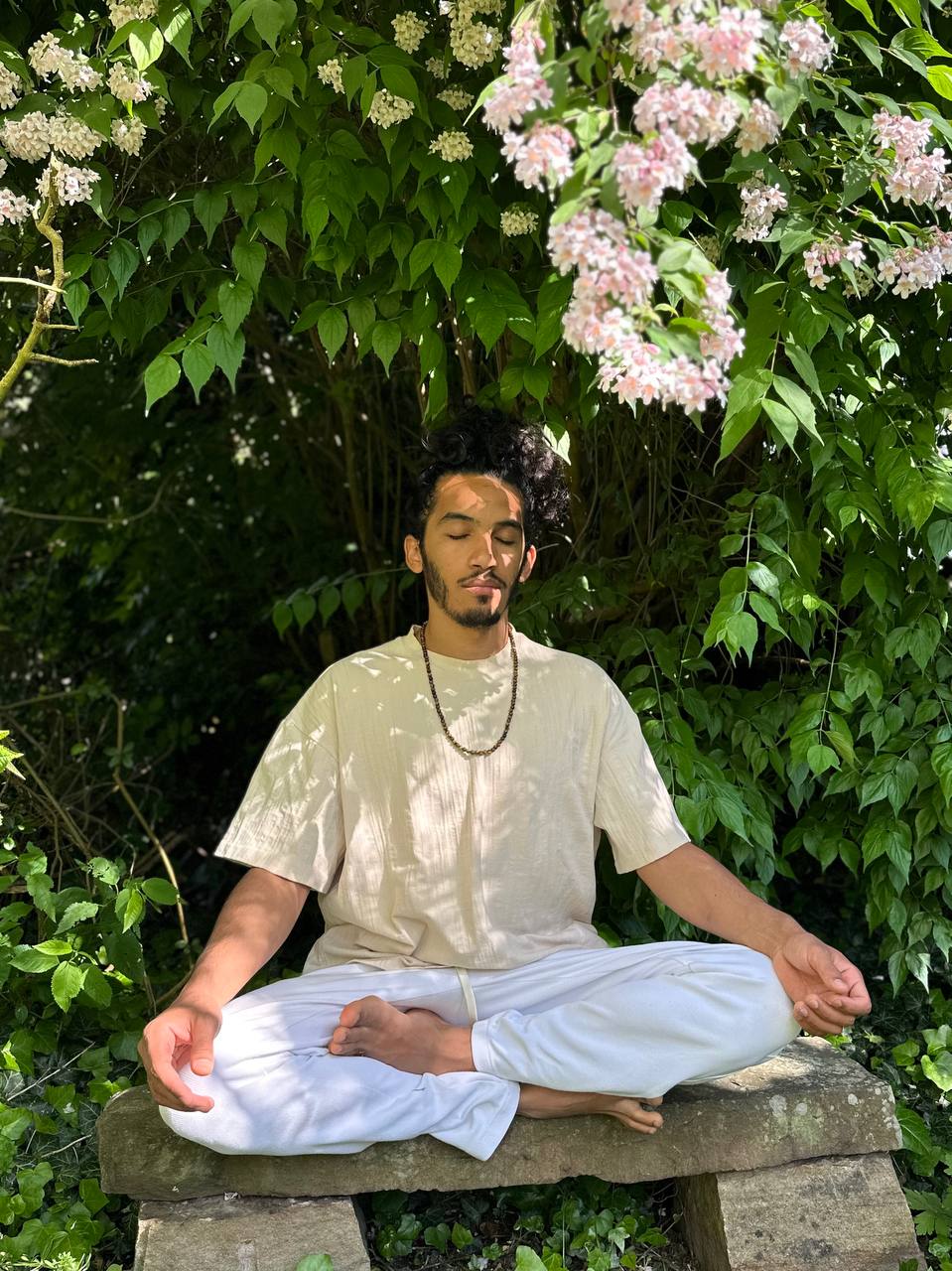 Lucas Santos meditando sentado em posição de lótus ao ar livre, cercado por vegetação e flores