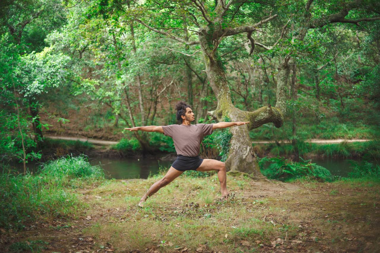 Lucas Santos praticando ioga ao ar livre em um bosque, em pé em uma postura de guerreiro com os braços estendidos